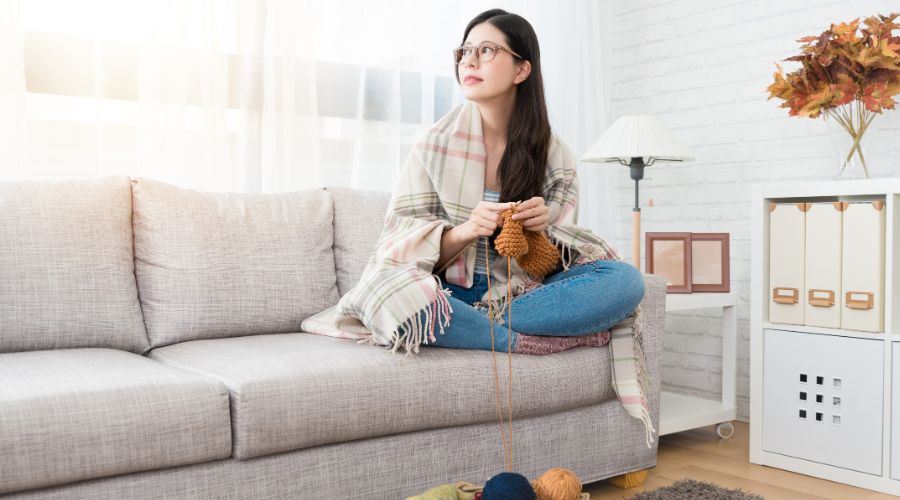 Woman knitting on a couch wrapped in a blanket, enjoying a cozy indoor setting, highlighting comfort during winter while managing heating costs.